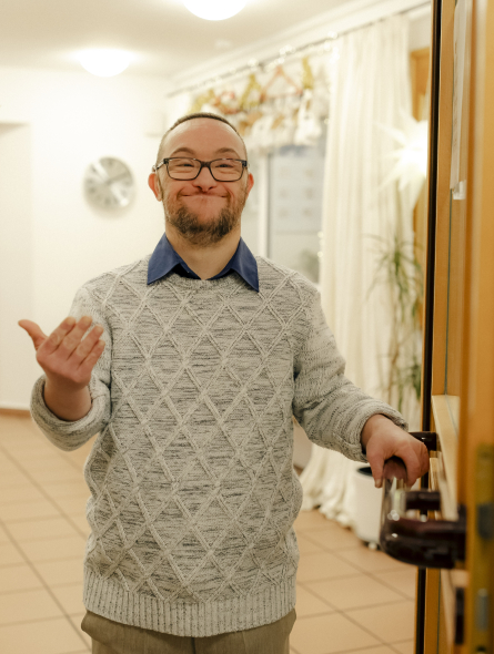 Ein Mann mit Behinderung steht in einer geöffneten Tür und winkt herein.