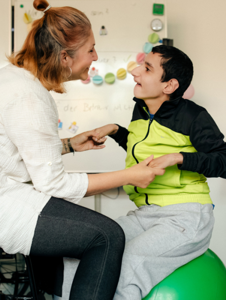 Eine Frau spricht mit einem Jungen, der auf einem Gymnastikball sitzt. Beide schauen sich an.