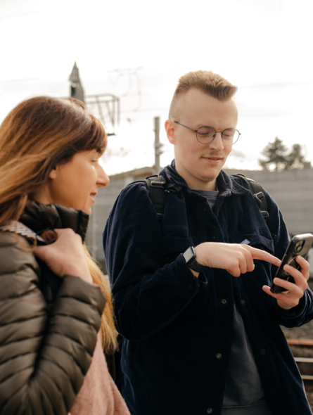 Eine Frau und ein junger Mann stehen am Bahnhof und schauen in ein Handy