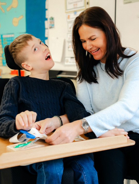 Eine Frau betreut und unterstützt einen Jungen, der im Rollstuhl sitzt, in der Schule.