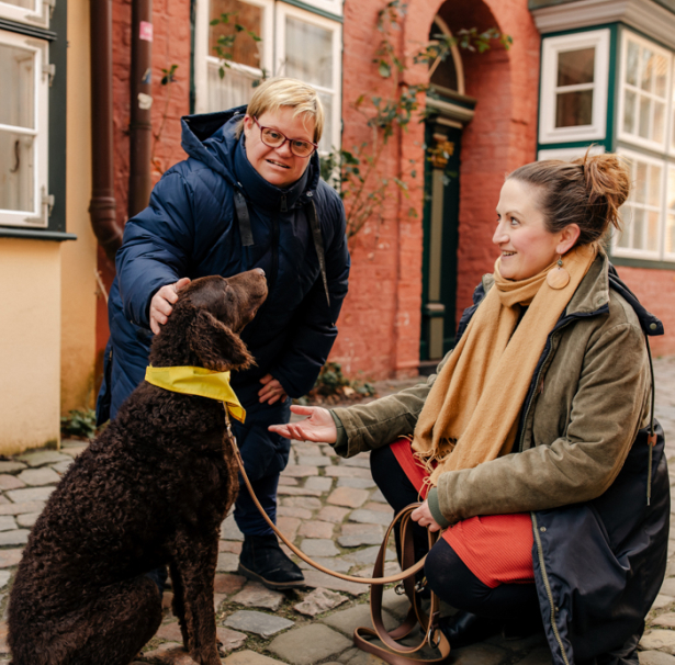 Eine Frau mit Behinderung streichelt einen Hund, den eine Frau an der Leine hat.