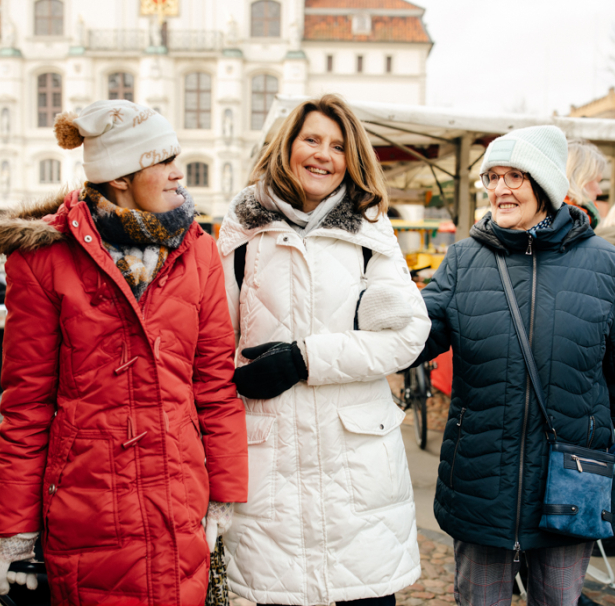 3 Frauen sind auf dem Wochenmarkt. Sie lächeln sich an. Im Hintergrund sieht man das Rathaus Lüneburg. 