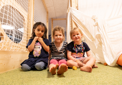 Drei Kleinkinder sitzen auf einer grünen Matte nebeneinander und schauen in die Kamera.