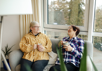 Zwei Seniorinnen mit Behinderung sitzen in Sesseln im Wintergarten und unterhalten sich.