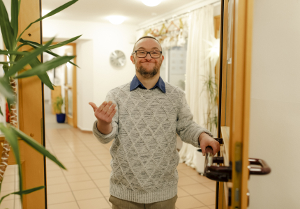Ein Mann mit Behinderung steht in einer geöffneten Tür und winkt herein.