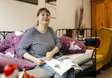 Eine junge Frau mit Behinderung sitzt auf ihrem Bett in ihrem Zimmer mit einer Zeitung auf dem Schoß.