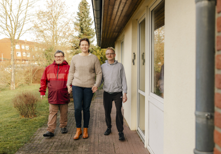 Eine Frau geht mit zwei Männern mit Behinderung an der Hand an der Außenwand eines Hauses vorbei.