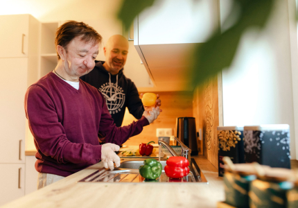 Ein Mann mit Behinderung reicht einem Mann hinter ihm eine Paprika. Beide stehen in der Küche am Herd.