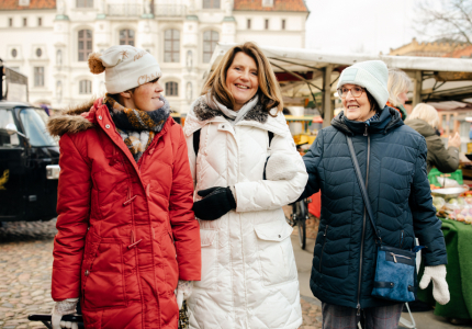 Drei Frauen mit und ohne Behinderung in Wintermänteln gehen über den Markt. 