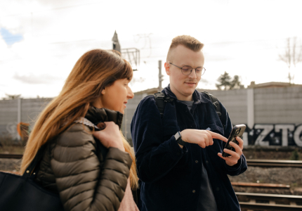 Eine Frau und ein junger Mann stehen am Bahnhof und schauen in ein Handy