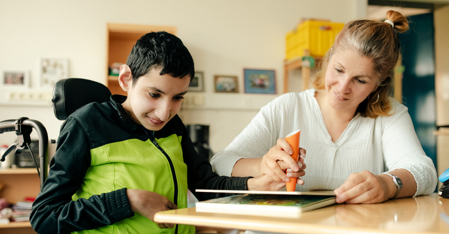 Eine Schulassistenz sitzt mit einem Jungen an einem Tisch und arbeitet gemeinsam in einem Buch.
