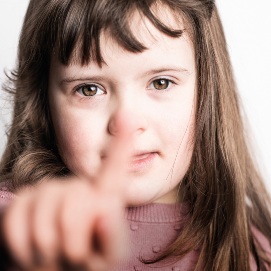 Ein junges Mädchen mit Trisomie 21 zeigt mit dem Finger in die Kamera