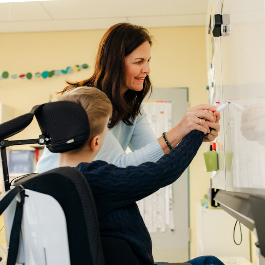 Eine Frau unterstützt einen Jungen, der im Rollstuhl sitzt, dabei, etwas an eine Tafel zu schreiben.
